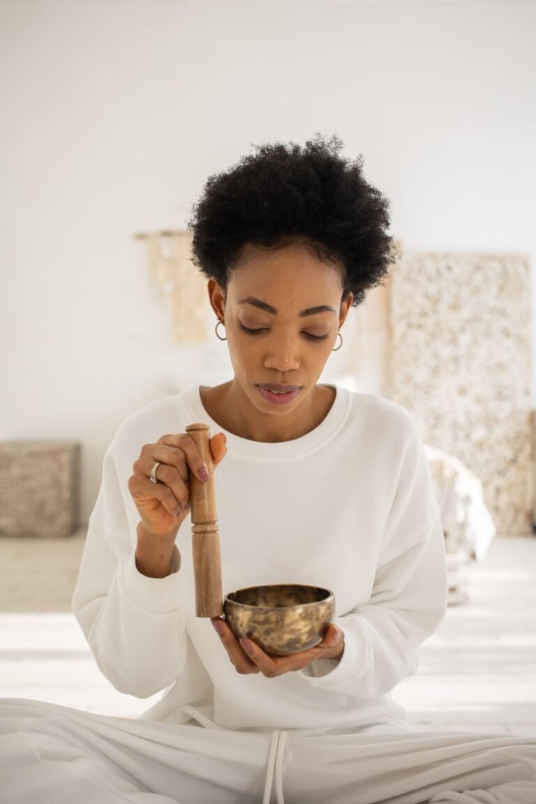 A woman in white practices meditation with a Tibetan singing bowl indoors.